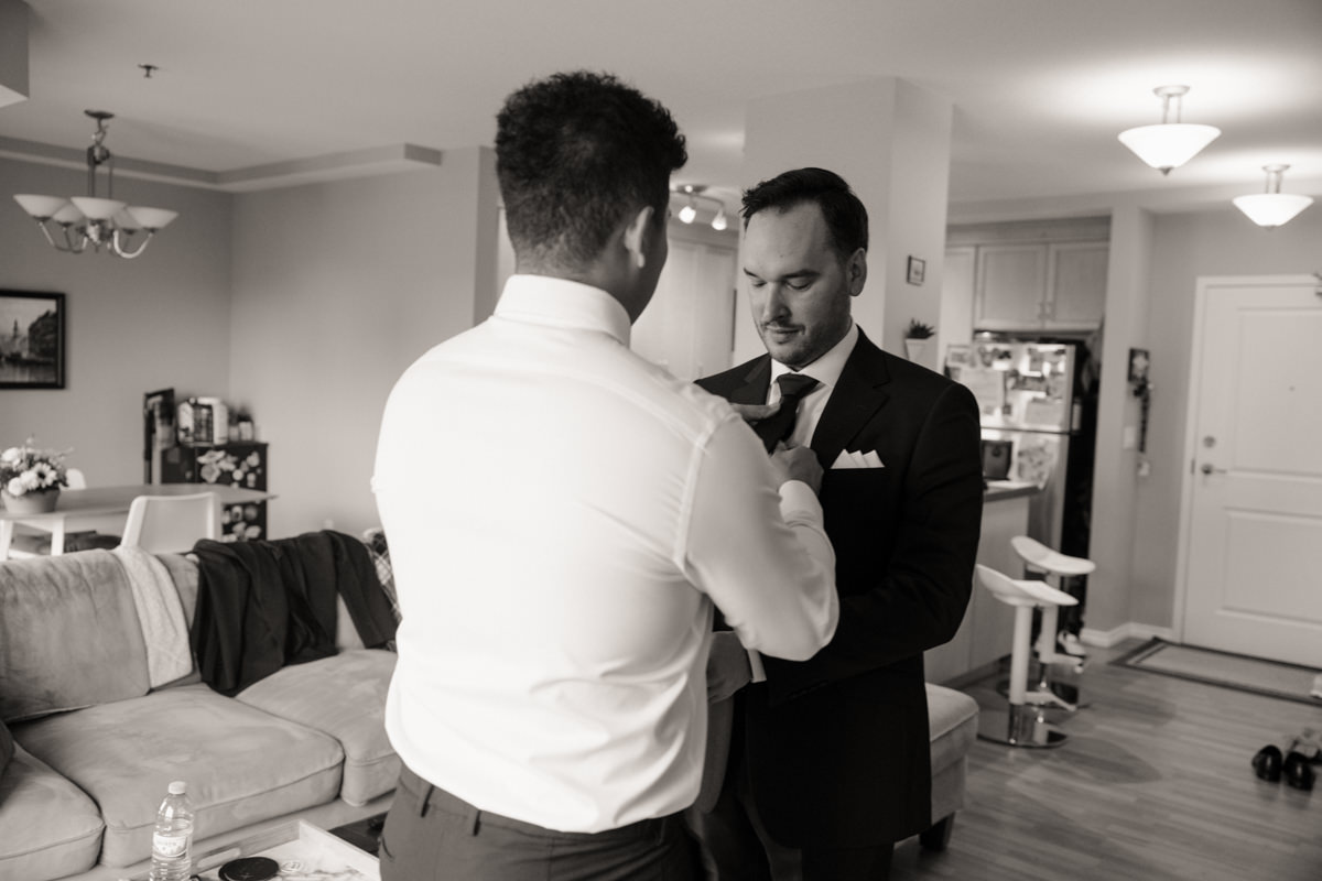 Grooman adjusting his tie while getting ready in a classic black tux at the Sheraton, preparing for his timeless Saskatoon wedding.