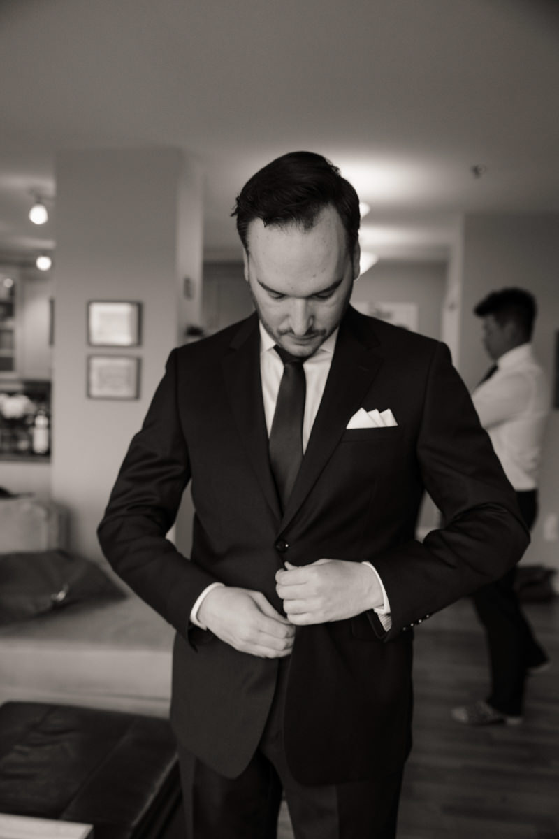Groom getting ready in a classic black suit, tying his tie before an elegant Saskatoon wedding ceremony