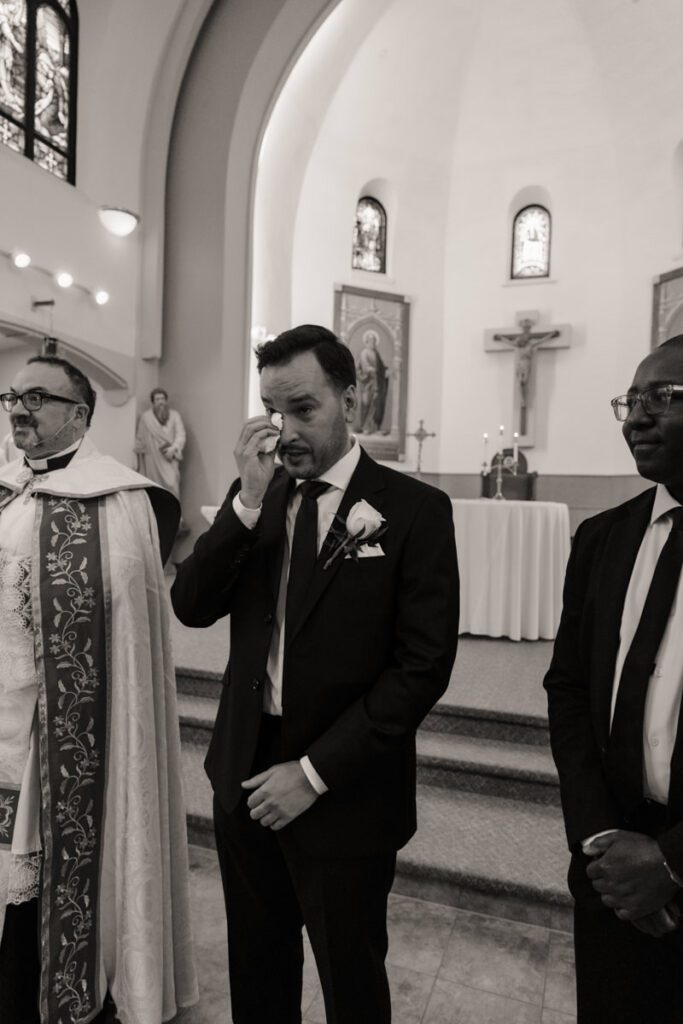 Groom watching his bride with emotion as she enters the cathedral, sunlight pouring through the stained glass windows.
