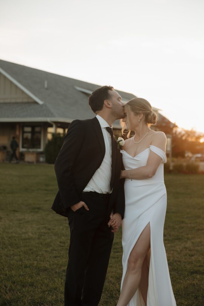 Silhouette of the newlyweds sharing a kiss at sunset, framed by trees and golden light during their elegant fall wedding.